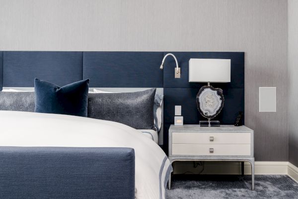 A modern bedroom featuring a blue and gray color scheme, with a bed, pillows, nightstand, lamp, and decorative items on it, all on a plush carpet.