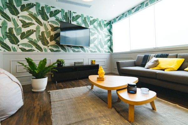 A living room with a tropical theme, featuring a green leaf wallpaper, gray sofa with yellow pillows, a wooden coffee table, and a large TV.