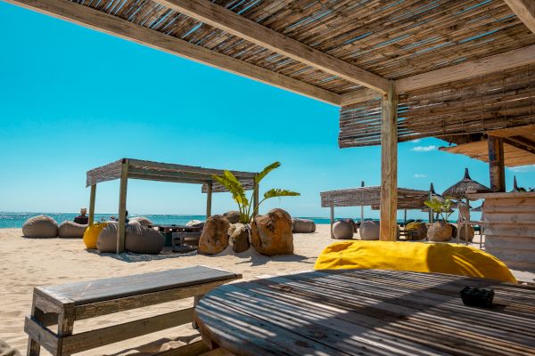 A beachfront setting with shaded seating areas, wooden tables, bean bags, and thatched umbrellas under a clear blue sky.