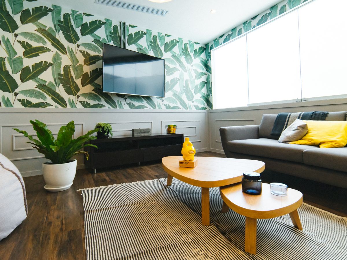A modern living room with tropical wallpaper, a wall-mounted TV, a gray couch with yellow cushions, a wooden coffee table, plants, and a rug.