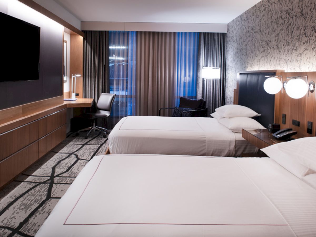 This image shows a modern hotel room with two neatly made beds, a flat-screen TV, a work desk and chair, and stylish decor with patterned wallpaper.