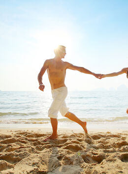 A couple holding hands runs along the beach, enjoying a sunny day with the ocean in the background.