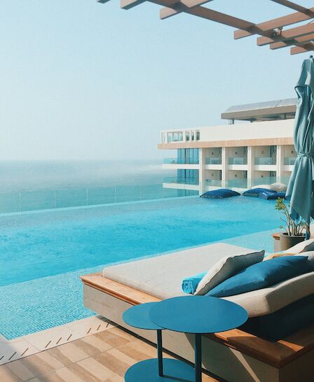 A luxurious poolside lounge area with cushioned seating, blue umbrellas, and a serene view of the ocean, located at a modern resort.