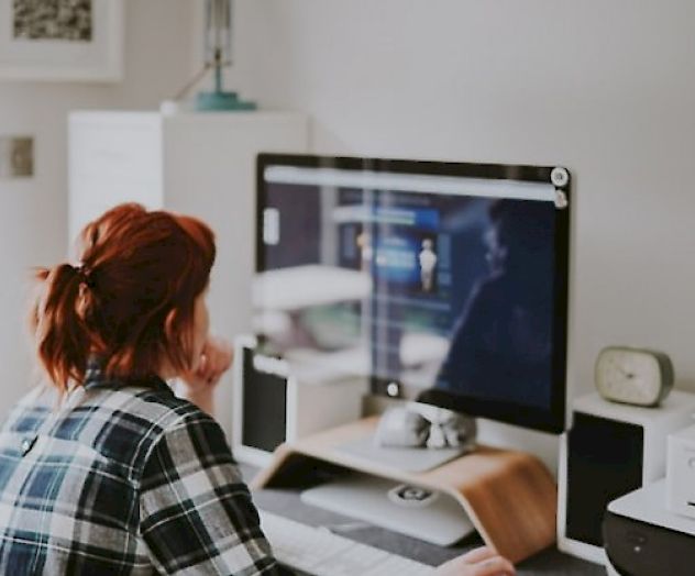 A person with red hair is sitting at a desk working on a computer, with various office supplies and gadgets around them.