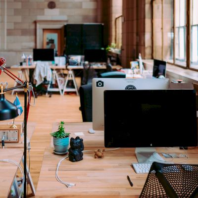 An open office space with wooden desks, computer screens, lamps, plants, and window lighting. There are chairs and various office supplies on desks.