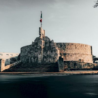 An image of a large stone monument with steps leading up to it, featuring a flag on top and surrounded by an empty road.
