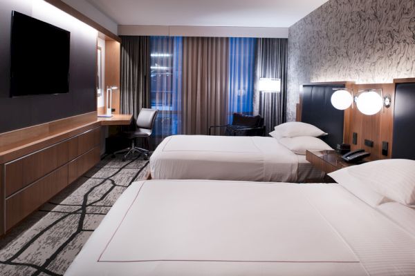 The image shows a modern hotel room with two beds, a large flat-screen TV, a desk with a chair, and contemporary decor and lighting.