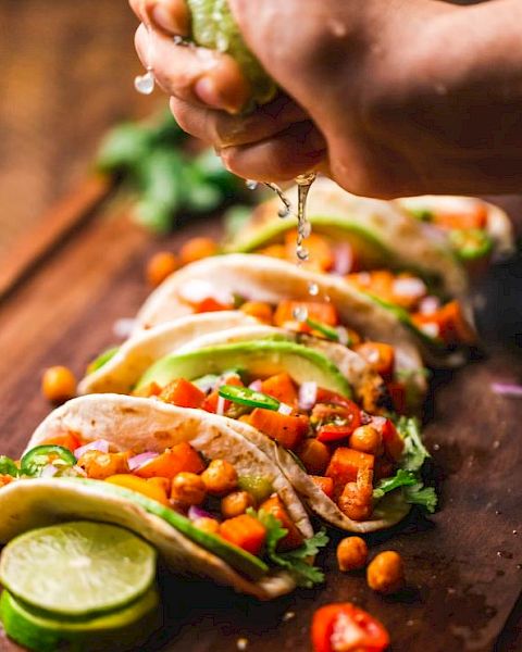 The image shows a hand squeezing lime juice over several tacos filled with chickpeas, diced tomatoes, avocado slices, red onions, and cilantro.