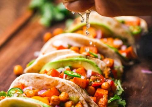 The image shows a hand squeezing lime juice over several tacos filled with chickpeas, diced tomatoes, avocado slices, red onions, and cilantro.