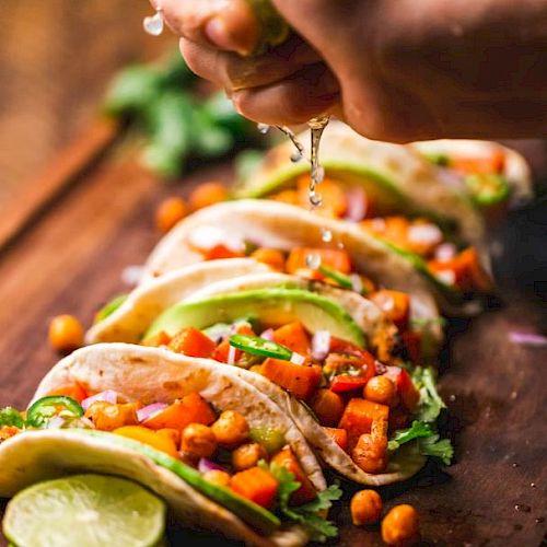 The image shows a hand squeezing lime juice over several tacos filled with chickpeas, diced tomatoes, avocado slices, red onions, and cilantro.
