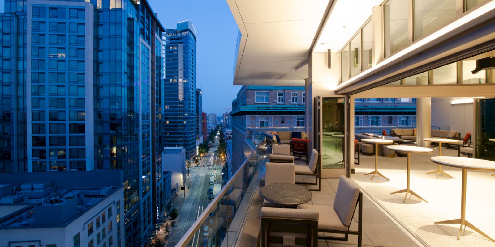 An outdoor balcony with seating and tables overlooks a cityscape at dusk, with tall buildings and illuminated streets in the background.