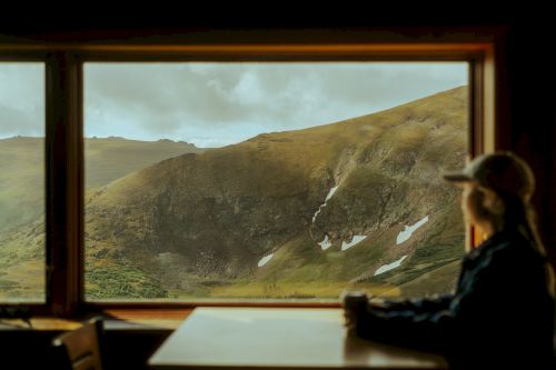 A person wearing a hat sits at a table, gazing out a window at a scenic mountain landscape with patches of snow.