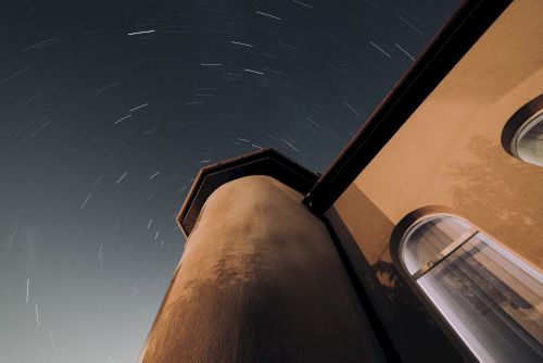 The image shows a night sky with star trails around a tall, rounded building structure. The building has arched windows on the right side.