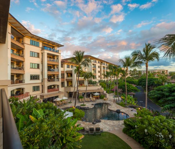 The image shows a tropical resort with multi-story buildings, a swimming pool, palm trees, lush greenery, and a partly cloudy sky.