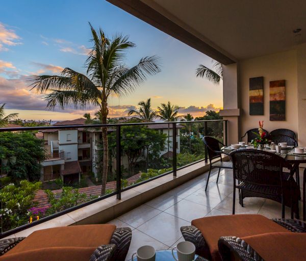 A tropical balcony features a dining table with chairs, overlooking lush greenery and palm trees against a colorful sunset sky.