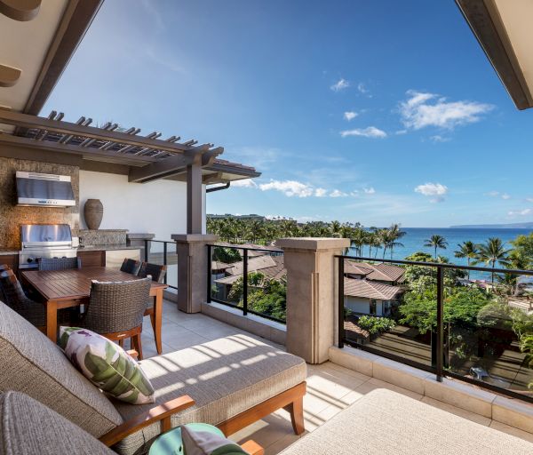 A luxurious terrace with lounge chairs, a dining area, and a grill overlooks a serene coastal view with palm trees and clear blue skies.