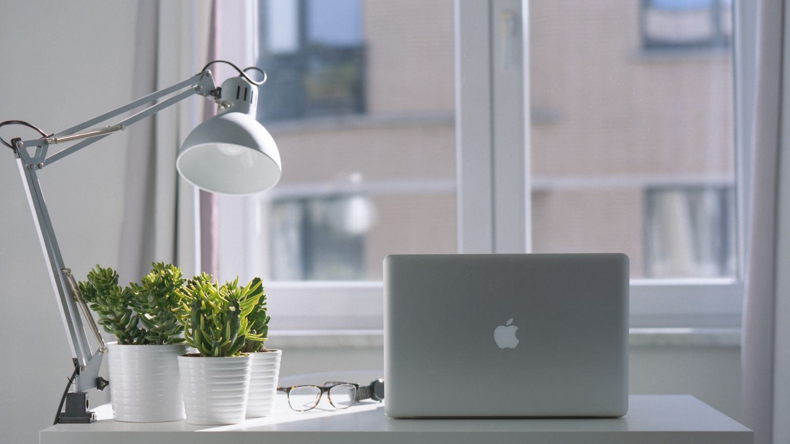 A minimalist desk setup with a laptop, potted plants, lamp, and eyeglasses, positioned in front of a window with curtains in a bright room.