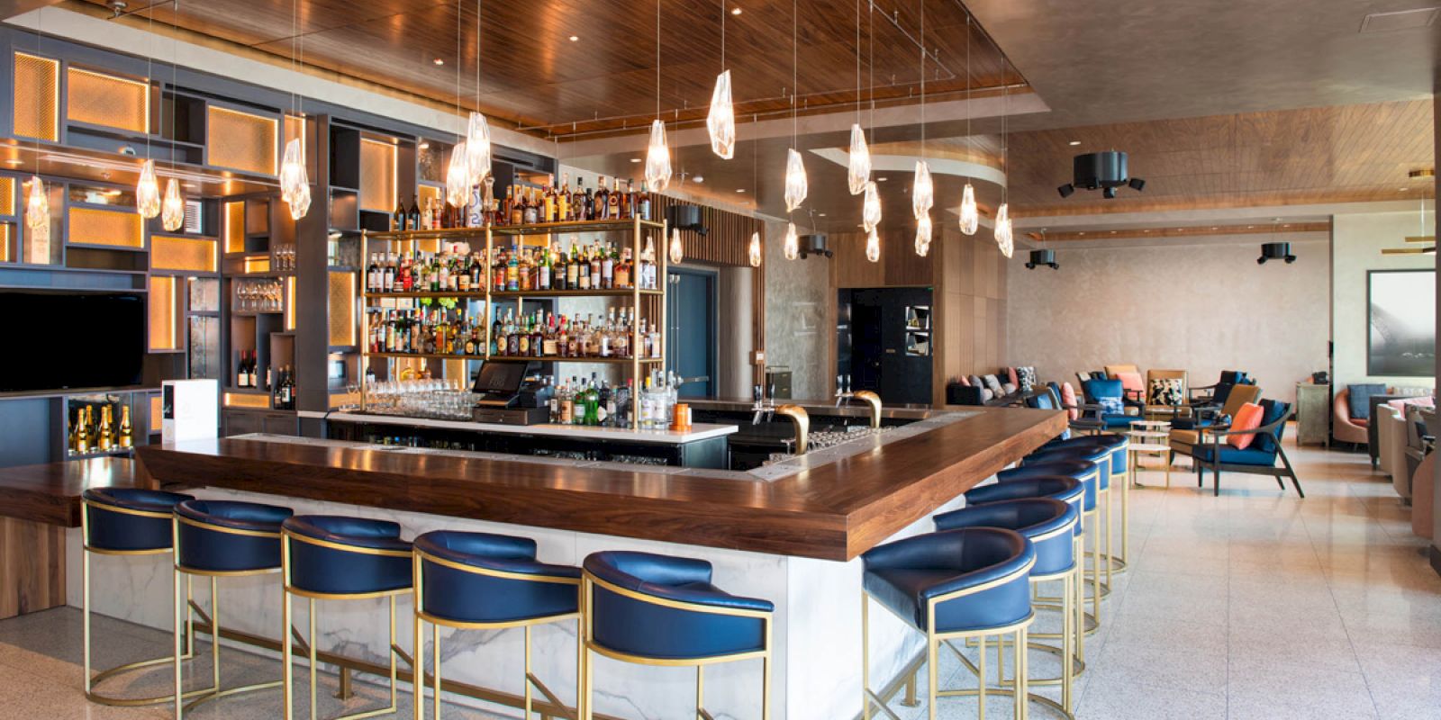 This image shows a modern bar with a central counter surrounded by blue and gold stools, stocked shelves of liquor, and hanging pendant lights.