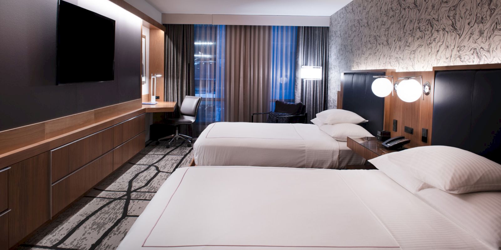 The image shows a modern hotel room with two double beds, a large flat-screen TV, a desk with a chair, and contemporary lighting and decor.