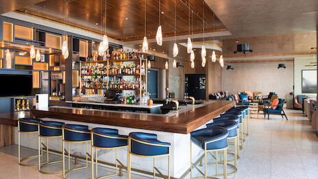 A modern bar with a well-stocked counter, surrounded by blue high chairs and hanging lights, with additional seating in the background.