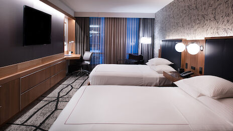 The image showcases a modern hotel room with two neatly made beds, a wall-mounted TV, a wooden desk with a chair, and patterned carpeting.