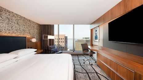 This image shows a modern hotel room with a large bed, a wall-mounted TV, a desk, and a city view through large windows.