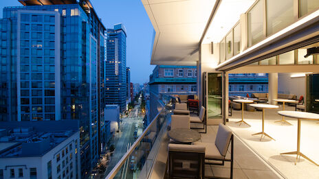 This image shows a modern city view from a high-rise balcony with tables, chairs, and part of an indoor area, all lit up in the evening.