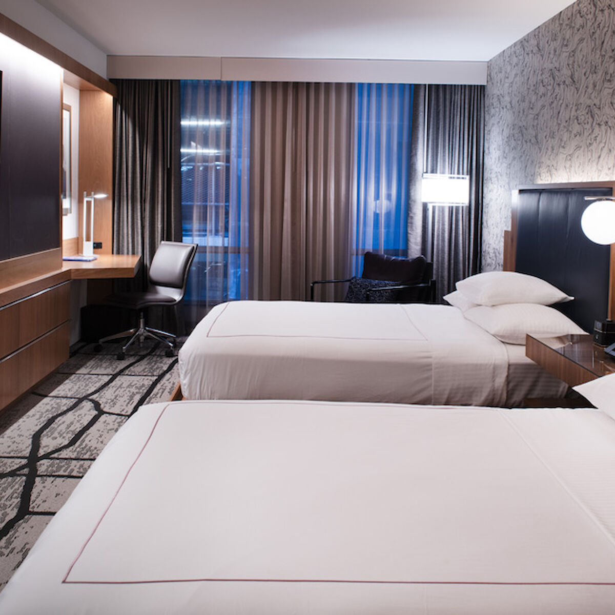 The image shows a modern hotel room with two neatly made beds, a large TV, a desk with a chair, and contemporary decor including wall lamps and patterned wallpaper.