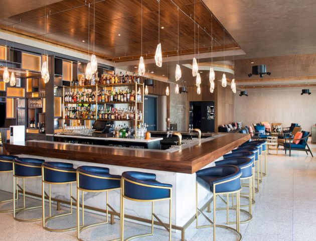 A modern bar with a wooden counter, high stools, pendant lights, and a well-stocked shelf, with seating areas in the background.