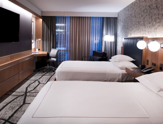 A hotel room with two beds, modern decor, a flat-screen TV, a desk with a chair, bedside lamps, and large windows with curtains.