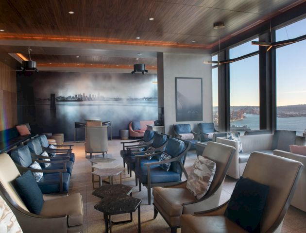 A modern lounge with numerous chairs, a panoramic view of the city and waterfront, a mural of a city skyline, and ambient lighting.