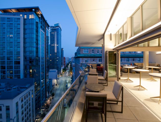 The image shows a modern cityscape with high-rise buildings and a spacious outdoor terrace at dusk, featuring tables and chairs for seating.
