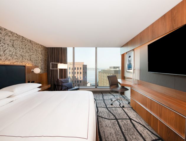 A modern hotel room features a large bed, a wall-mounted TV, a desk with a chair, and a window with a city view.