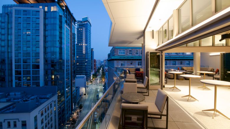 The image shows a modern balcony overlooking a city street filled with tall buildings at dusk. The balcony has tables and chairs.