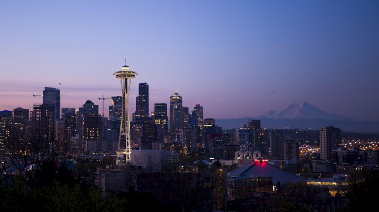 A twilight cityscape featuring a tall structure with a spire, various buildings, and a mountain in the background under a purple and blue sky.