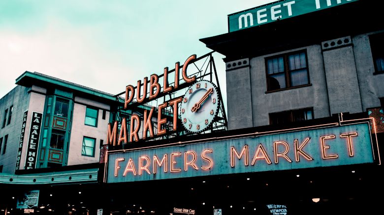 An outdoor scene featuring a neon sign reading "Public Market" and "Farmers Market" with buildings in the background and a clock integrated into the sign.